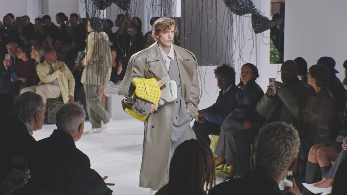 Sebuah jas panjang berwarna cerah dengan potongan bahu yang tegas, tambahan aksesori tas tangan kuning menjadi fokus utama.